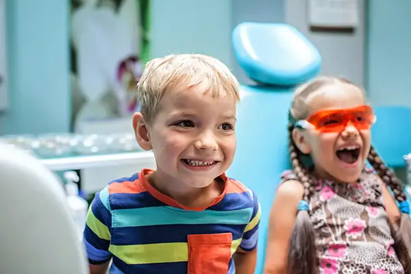 Two giddy children sitting in the dentist chair and smiling with one playfully wearing protective orange glasses.